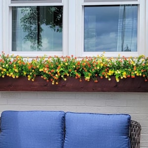 May include: A wooden window box planter filled with yellow and orange flowers. The planter is mounted below a white window with two panes.