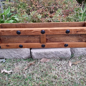 May include: A wooden planter box with a stained brown finish and black metal accents. The planter has a rectangular shape and is sitting on a stone surface.