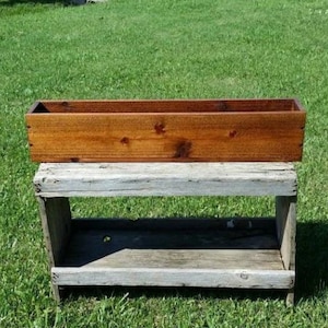 May include: A rectangular wooden planter box with a rich brown stain sits atop a weathered, gray wooden stand. The planter box is empty, and the scene is set outdoors on a bed of green grass.