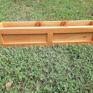 May include: A wooden planter box with a natural finish. The planter has a rectangular shape with a raised lip around the top and a single divider in the middle.