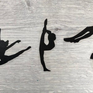 Op de afbeelding: Drie zwarte silhouetuitsparingen van gymnasten in verschillende poses. De figuren zijn geplaatst tegen een lichtgrijze houtnerf achtergrond. Eén gymnaste staat in een split-sprong, een ander in een verticale split en de derde in een rugbuiging.