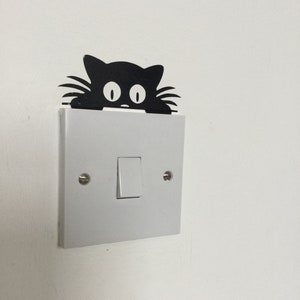 May include: A white light switch with a black cat sticker peeking over the top. The cat has large, round eyes.