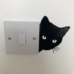 Könnte beinhalten: Ein weißer Lichtschalter mit einem schwarzen Katzenaufkleber, der hinter ihm hervorlugt. Die Katze hat weiße Augen und Schnurrhaare.