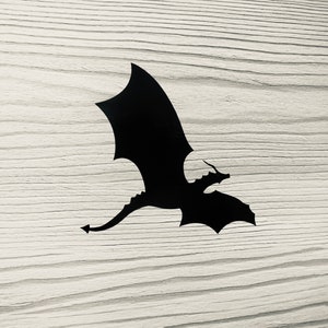 Può includere: Silhouette nera di un drago con le ali spiegate, che vola su uno sfondo di legno chiaro.