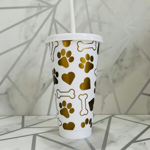 May include: White reusable cup with a gold paw print, bone, and heart pattern.