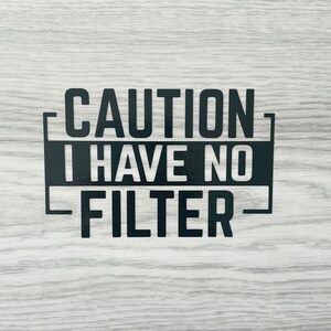Può includere: Un cartello bianco e nero con il testo "CAUTION I HAVE NO FILTER" su uno sfondo in legno.