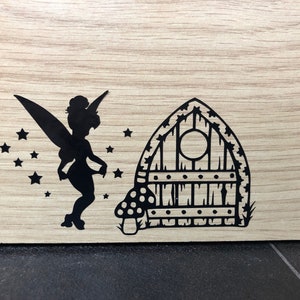 May include: Black silhouette of a fairy with wings and a star-shaped wand standing in front of a small, black, wooden door with a round window. The door is surrounded by a mushroom and grass.