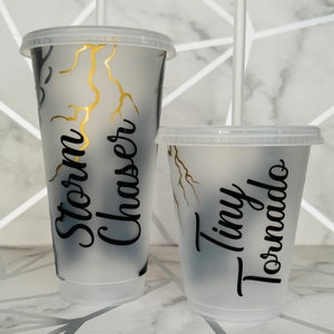 Peut inclure: Deux gobelets en plastique transparent avec des motifs d'éclairs noirs et dorés. Le plus grand gobelet dit "Storm Chaser" et le plus petit dit "Tiny Tornado".