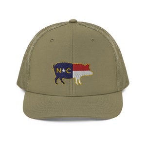 May include: Olive green trucker hat with a mesh back and a curved brim. The front of the hat features an embroidered pig design with the North Carolina state flag colors and the letters "N*C".