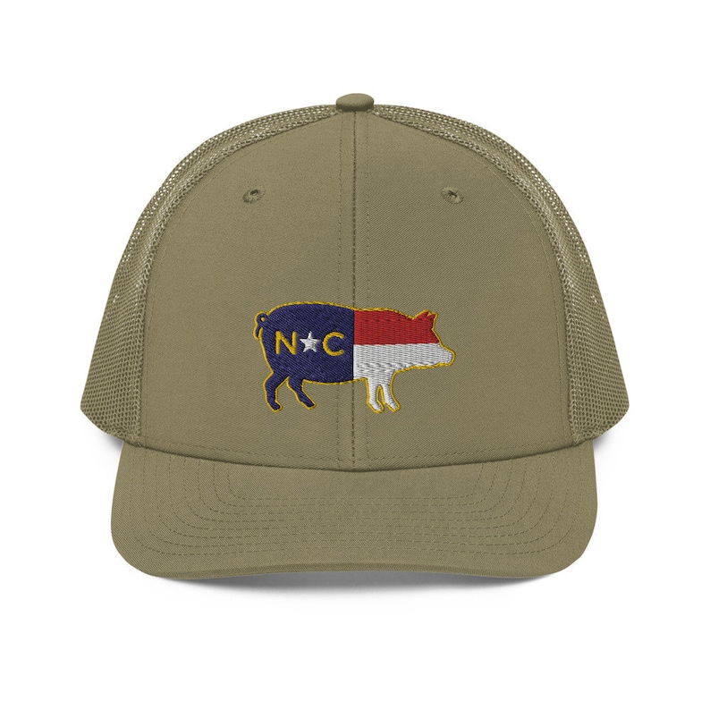 May include: Olive green trucker hat with a mesh back and a curved brim. The front of the hat features an embroidered pig design with the North Carolina state flag colors and the letters "N*C".