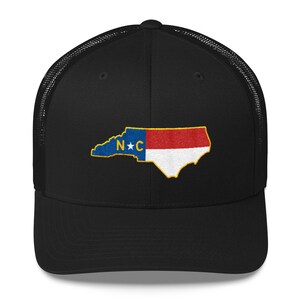 May include: Black trucker hat with a mesh back. The front of the hat features an embroidered outline of the state of North Carolina with the state flag colors and the letters "N*C" in the center.