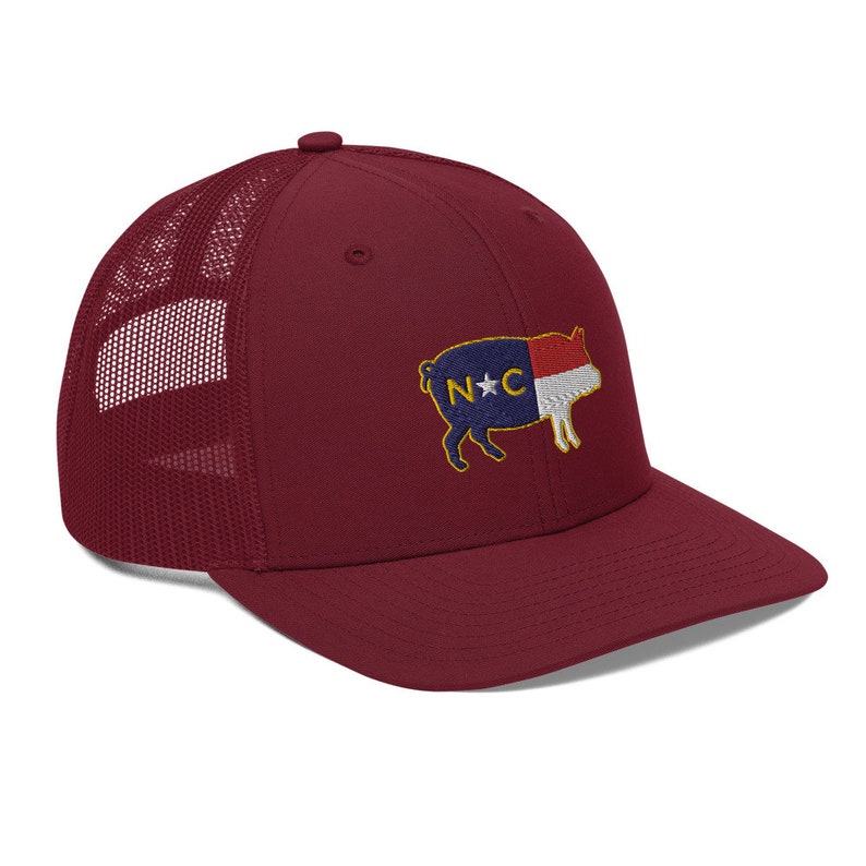 May include: A burgundy trucker hat with a mesh back and a curved brim. The hat features an embroidered patch of a pig with the North Carolina state flag design. The pig is in shades of blue, red, white, and gold.