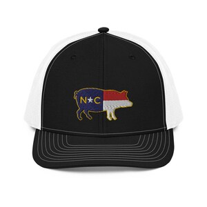 May include: Black and white trucker hat with a pig-shaped embroidered design. The pig features the North Carolina state flag colors and the letters "N" and "C". The hat has a curved brim and a mesh back.
