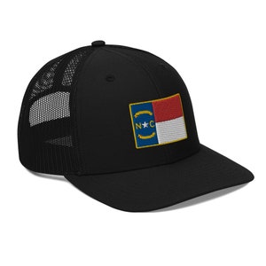 Op de afbeelding: Zwarte truckercap met een mesh-rug en een geborduurde vlag van de staat North Carolina aan de voorkant. De vlag is blauw, rood en wit met een gele ster en de letters "N.C."