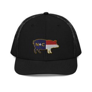 May include: Black trucker hat with a mesh back and a structured front. The hat features an embroidered pig design with the North Carolina state flag colors and the letters "N*C".