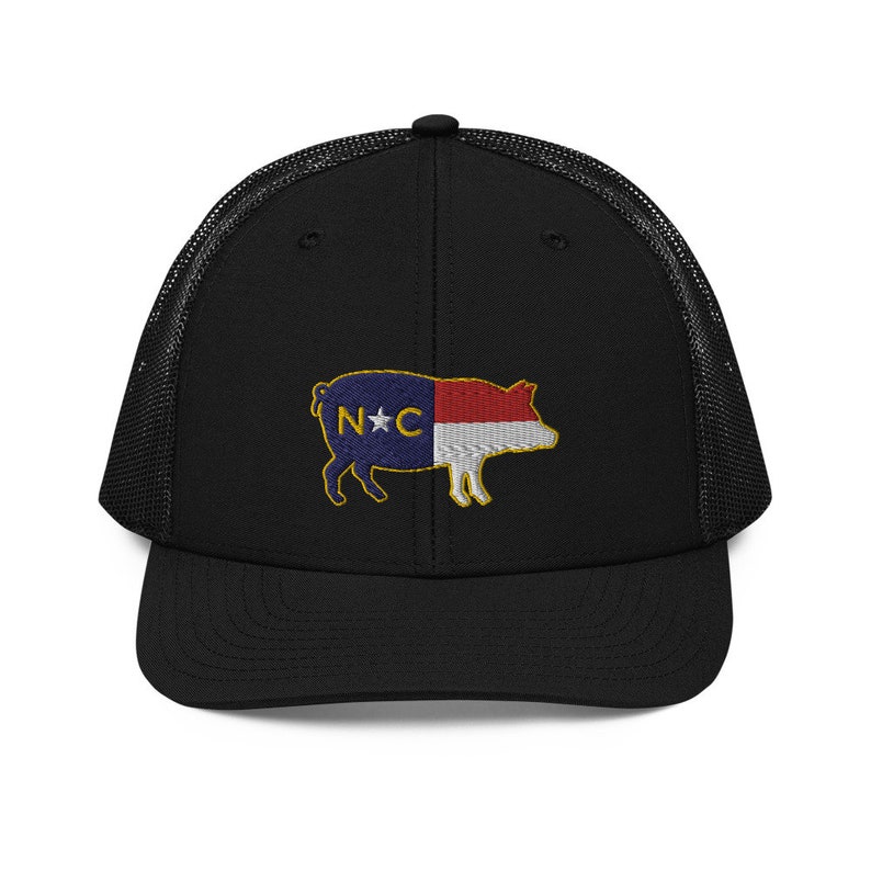May include: Black trucker hat with a mesh back and a structured front. The hat features an embroidered pig design with the North Carolina state flag colors and the letters "N*C".