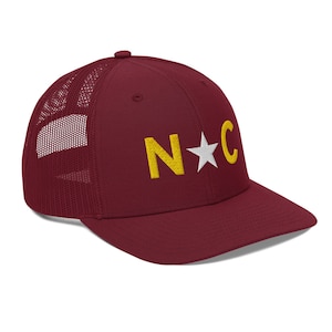 May include: A maroon trucker hat with a white mesh back. The front of the hat has a gold embroidered "N*C" design.