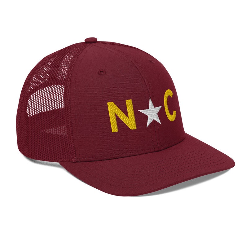 May include: A maroon trucker hat with a white mesh back. The front of the hat has a gold embroidered "N*C" design.