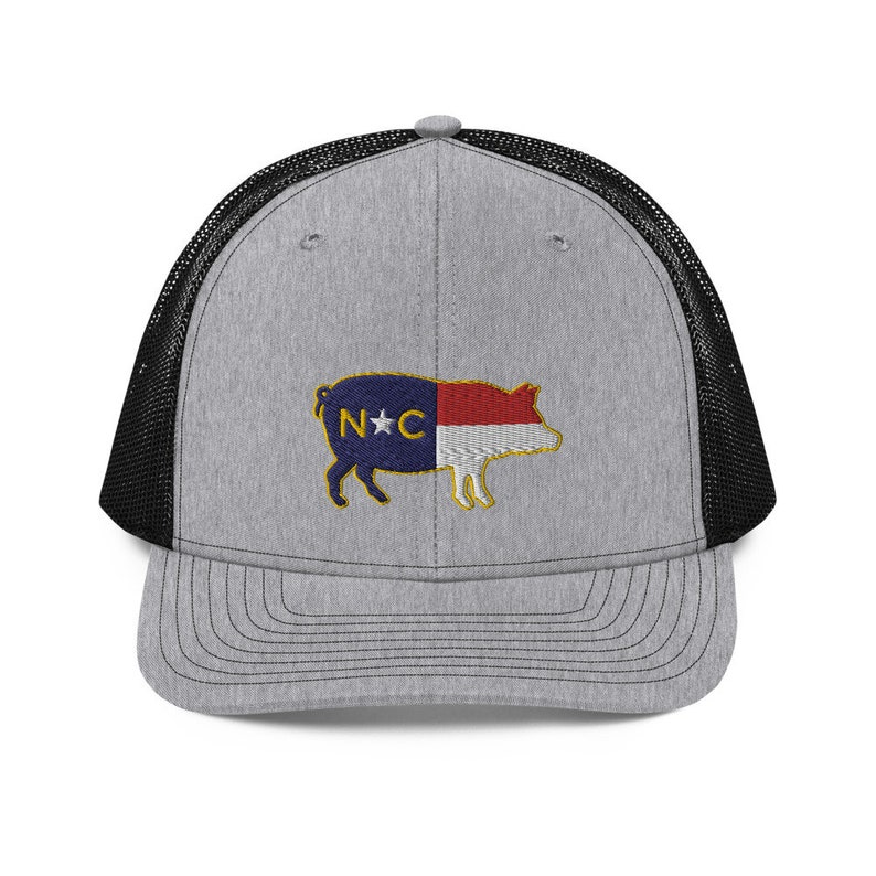 May include: Gray trucker hat with a black mesh back and a curved brim. The front features an embroidered pig design with the North Carolina state flag colors and the letters "NC".