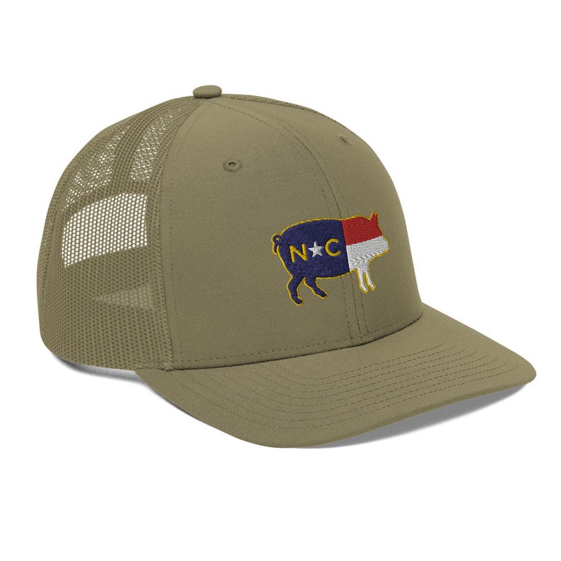 May include: Olive green trucker hat with a mesh back and a curved brim. The hat features an embroidered design of a pig with the North Carolina state flag colors and the letters "N*C".