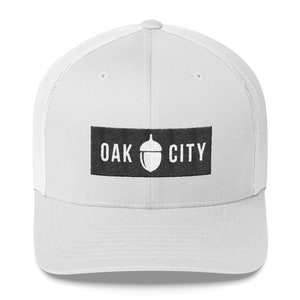 Może przedstawiać: Biała siatkowa czapka trucker z czarnym naszywką z napisem "OAK CITY" i grafiką żołędzia w środku.