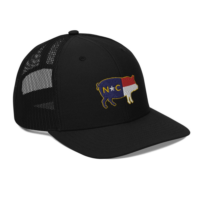 May include: Black trucker hat with a mesh back and a curved brim. The hat features an embroidered patch of a pig with the North Carolina state flag design. The pig is outlined in yellow, with the letters "N*C" on its side.