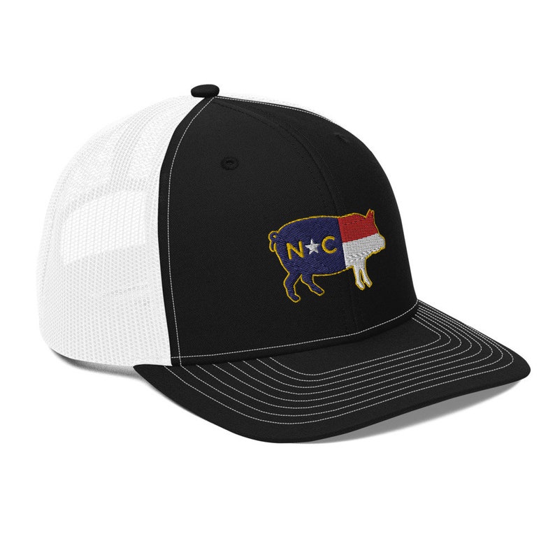 May include: Black and white trucker hat with a curved brim. The front panel is black, and the back is white mesh. An embroidered patch of a pig with the North Carolina state flag design is on the front.