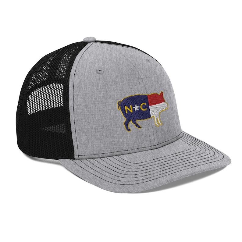 May include: A gray trucker hat with a black mesh back. The front of the hat features an embroidered pig design with the North Carolina state flag colors and the letters "N" and "C". The hat has a curved brim and a snapback closure.
