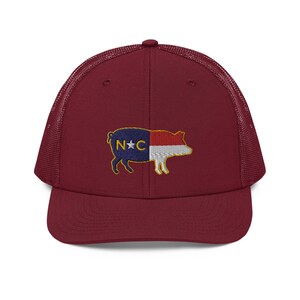 May include: A burgundy trucker hat with a mesh back and a curved brim. The front of the hat features an embroidered pig design with the North Carolina state flag colors and the letters "N*C".