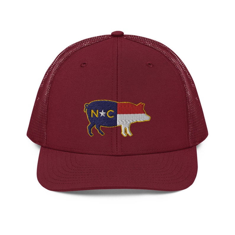 May include: A burgundy trucker hat with a mesh back and a curved brim. The front of the hat features an embroidered pig design with the North Carolina state flag colors and the letters "N*C".