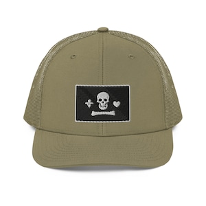 May include: A khaki green trucker hat with a black and white embroidered skull and crossbones patch. The patch features a heart and a symbol that looks like a cross with a circle around it.