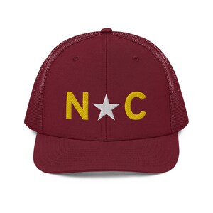 May include: A maroon trucker hat with a mesh back. The front of the hat features a white star and the letters "N*C" in yellow embroidery.