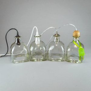 May include: Four clear glass bottle pendant lights with different tops and electrical cords. One has a black cord, one a white cord, one a silver top, and one a cork top with a green tag.
