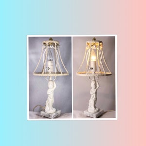 May include: A white table lamp with a white rope shade and a white cherub figurine base. The lamp is turned on and the light bulb is illuminated.