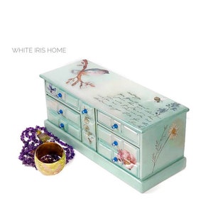 May include: A mint green jewelry box with multiple drawers and blue knobs. The box is decorated with floral and bird illustrations and script. A purple beaded necklace and a gold bracelet are next to the box. The text "WHITE IRIS HOME" is in the upper left.