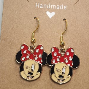 May include: A pair of gold-toned dangle earrings featuring a cartoon character design. Each earring showcases a smiling face with a red bow with white polka dots, black ears, and a yellow face. The earrings are on a brown card with the word "Handmade".