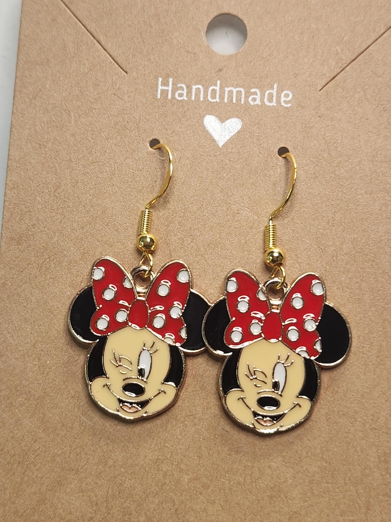 May include: A pair of gold-toned dangle earrings featuring a cartoon character design. Each earring showcases a smiling face with a red bow with white polka dots, black ears, and a yellow face. The earrings are on a brown card with the word "Handmade".