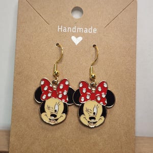 May include: A pair of gold-toned dangle earrings featuring Minnie Mouse. Each earring showcases Minnie's face with a red polka-dot bow, black ears, and a yellow face. The earrings are displayed on a brown card with the word "Handmade".