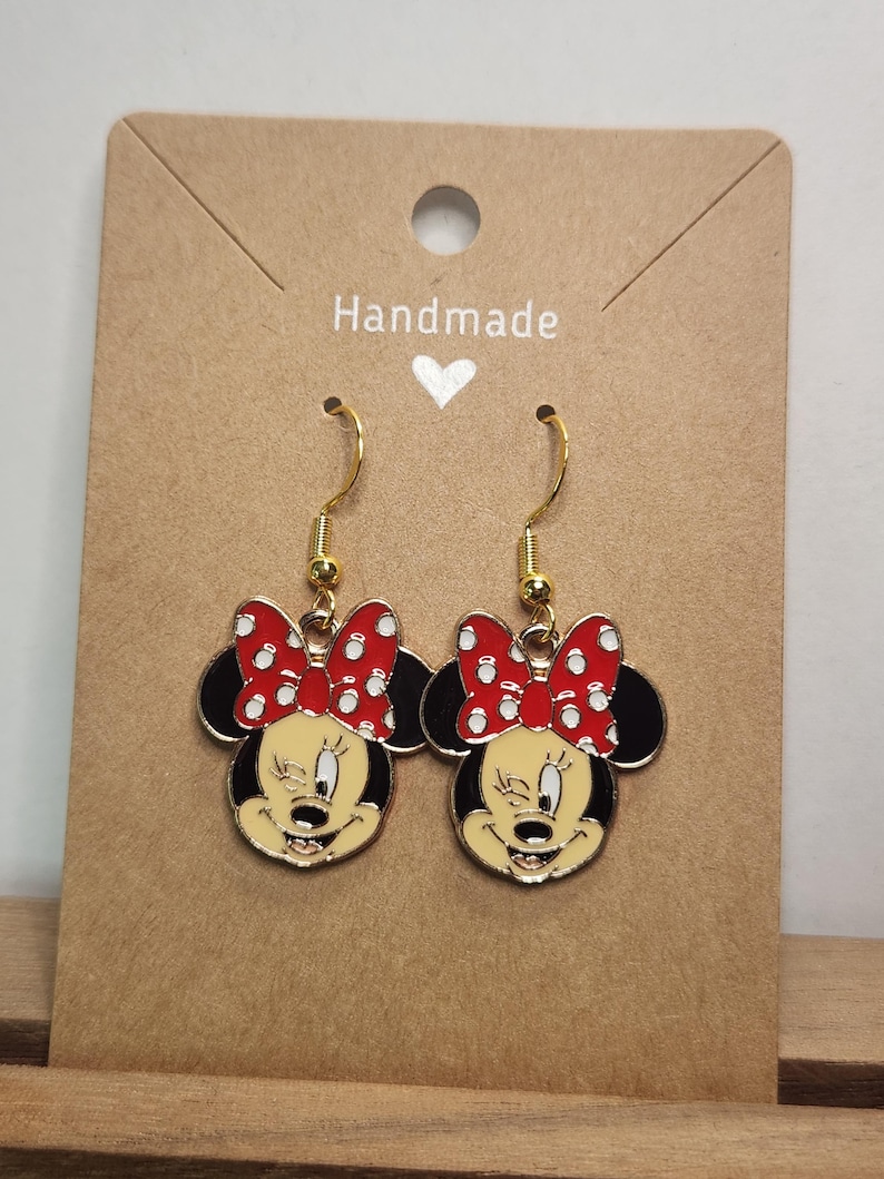 May include: A pair of gold-toned dangle earrings featuring Minnie Mouse. Each earring showcases Minnie's face with a red polka-dot bow, black ears, and a yellow face. The earrings are displayed on a brown card with the word "Handmade".