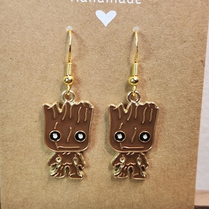 May include: A pair of gold-colored earrings featuring a brown cartoon character design with black and white details. The earrings are hanging on a brown card with the word "Handmade" and a white heart.