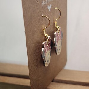 May include: Gold-tone Minnie Mouse dangle earrings. Each earring features a Minnie Mouse design with a red or pink bow, black ears, and a white face. The earrings are displayed on a brown card that reads "Handmade".