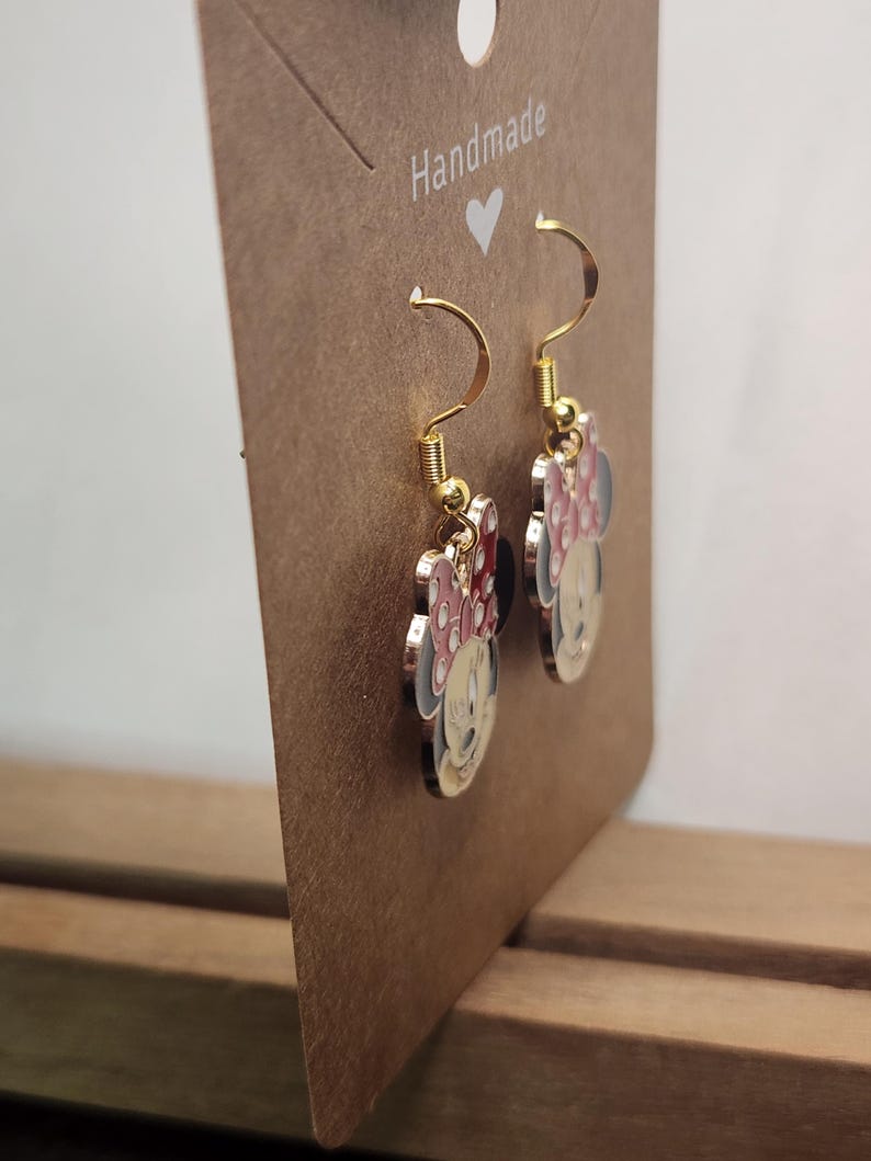 May include: Gold-tone Minnie Mouse dangle earrings. Each earring features a Minnie Mouse design with a red or pink bow, black ears, and a white face. The earrings are displayed on a brown card that reads "Handmade".