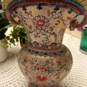 May include: A decorative ceramic vase with a gold background and floral patterns in red, blue, and pink. The vase has a flared top with handles and a round body. The design includes intricate details and a touch of elegance.