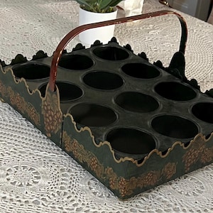 May include: A green metal bottle carrier with a brown handle. The carrier has 12 compartments and a decorative grape design.