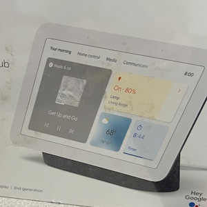 May include: A white smart display device with a dark gray base. The screen displays information including the time, weather, and music controls. The text "Hey Google" is visible on the packaging.