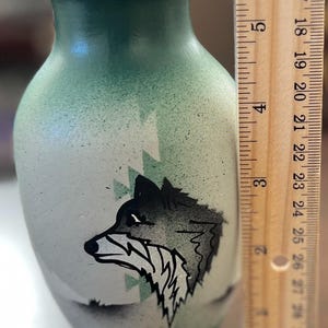 May include: A green and white vase featuring a black wolf head illustration and geometric patterns, positioned next to a wooden ruler showing the object is approximately 5 inches tall with a flared rim and a rounded body shape.