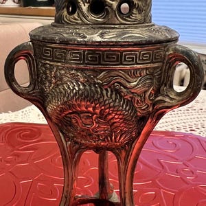 May include: Ornate silver-toned incense burner with a dragon motif. The burner has a domed lid with air holes and two handles. The base has three legs and sits on a red surface. The incense burner is a decorative item.