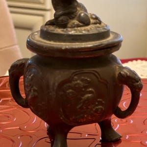 May include: A bronze-colored incense burner with a lid featuring a small, seated lion figure. The burner has ornate handles and decorative relief patterns. It stands on three short legs and is set against a red surface.