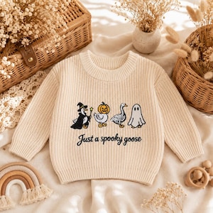 Puede incluir: Un suéter de punto de manga larga en color crema con un gráfico bordado de cuatro gansos con disfraces de Halloween, incluyendo una bruja, una cabeza de calabaza, un esqueleto y un fantasma, con el texto negro "Just a spooky goose".