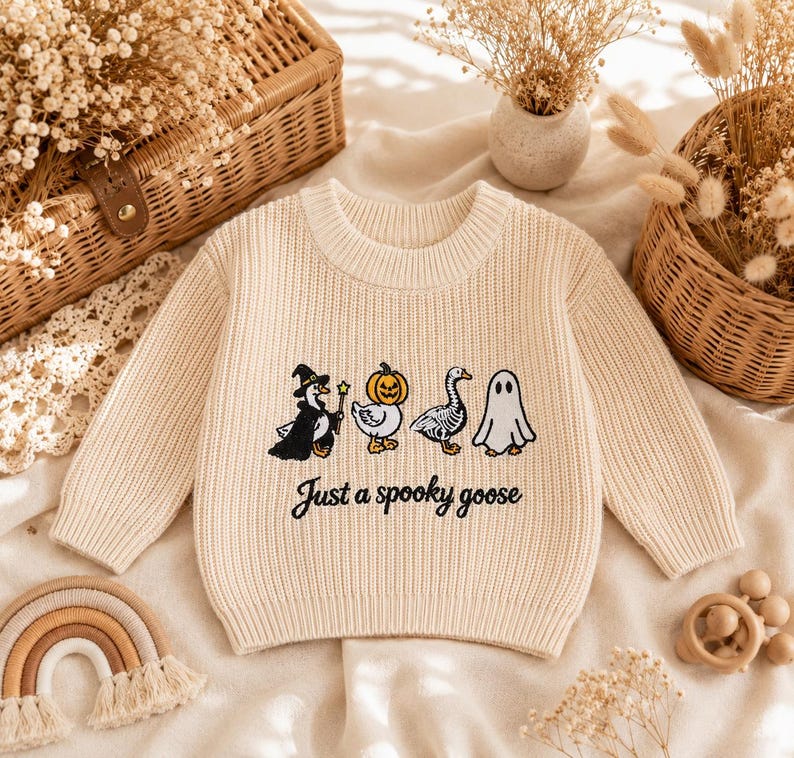 May include: A cream-colored long-sleeve knit sweater featuring an embroidered graphic of four geese in Halloween costumes, including a witch, a pumpkin head, a skeleton, and a ghost, with the black text "Just a spooky goose" printed below.